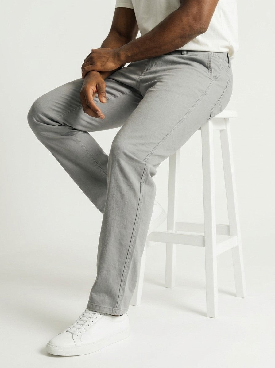 Homem sentado em banquinho usando calça chino cinza Bluviso com corte ajustado e tênis branco casual