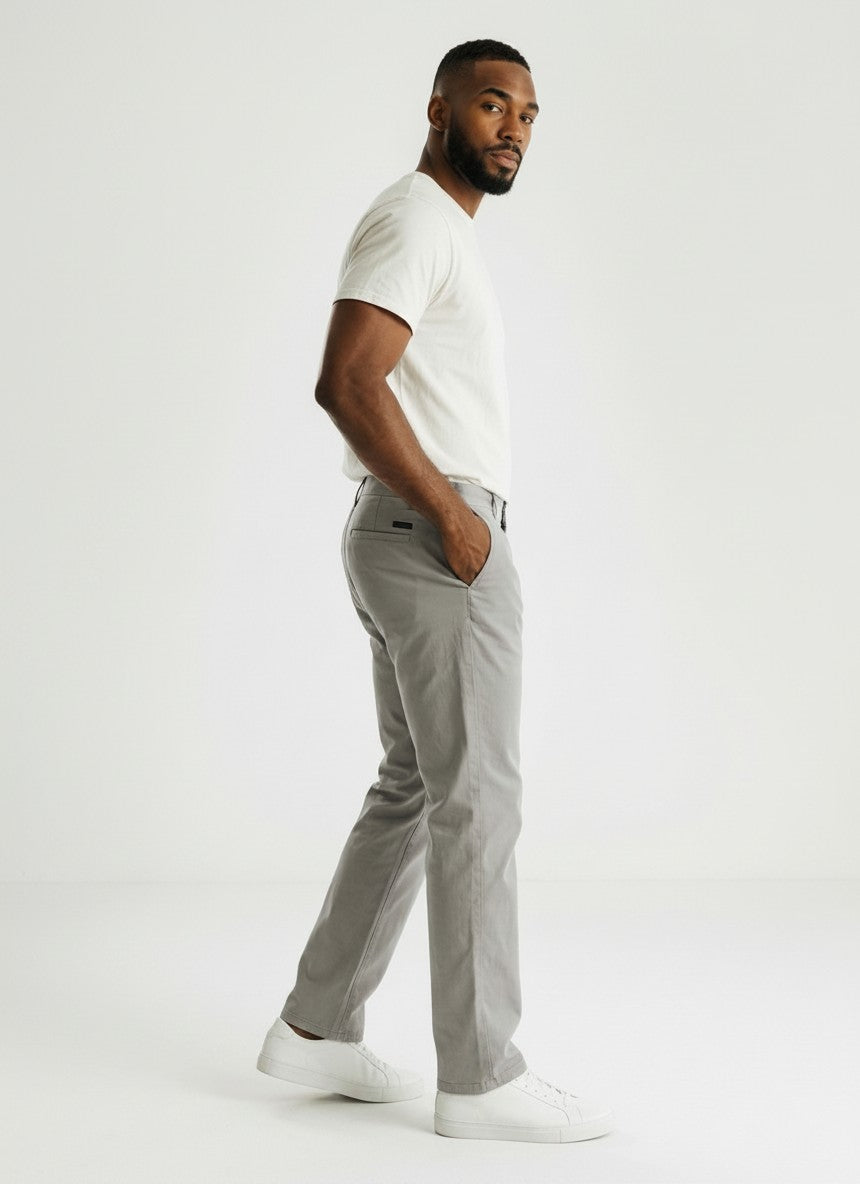 Homem vestindo calça chino cinza Bluviso com camiseta branca e tênis, vista lateral em estúdio com fundo neutro.