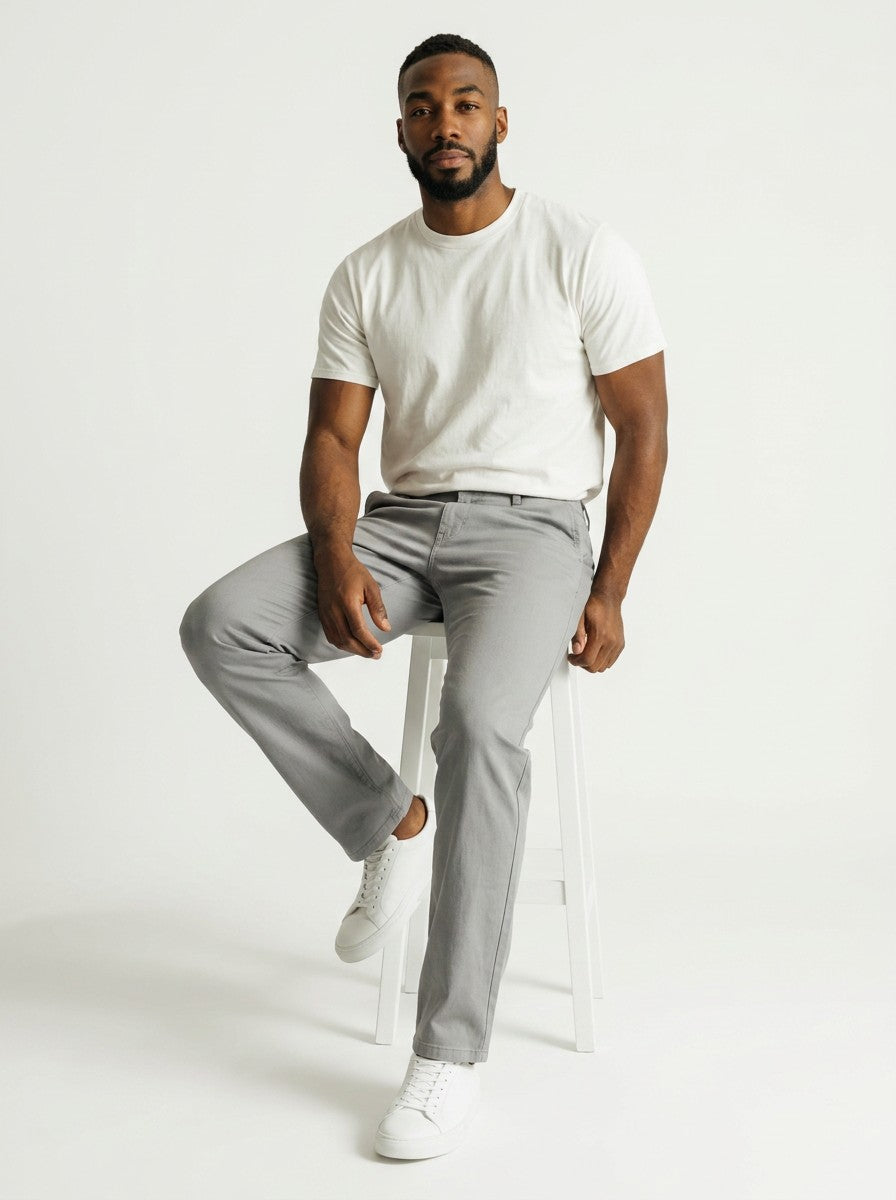Homem vestindo calça chino cinza Bluviso com camiseta branca e tênis, sentado em um banco contra fundo neutro.