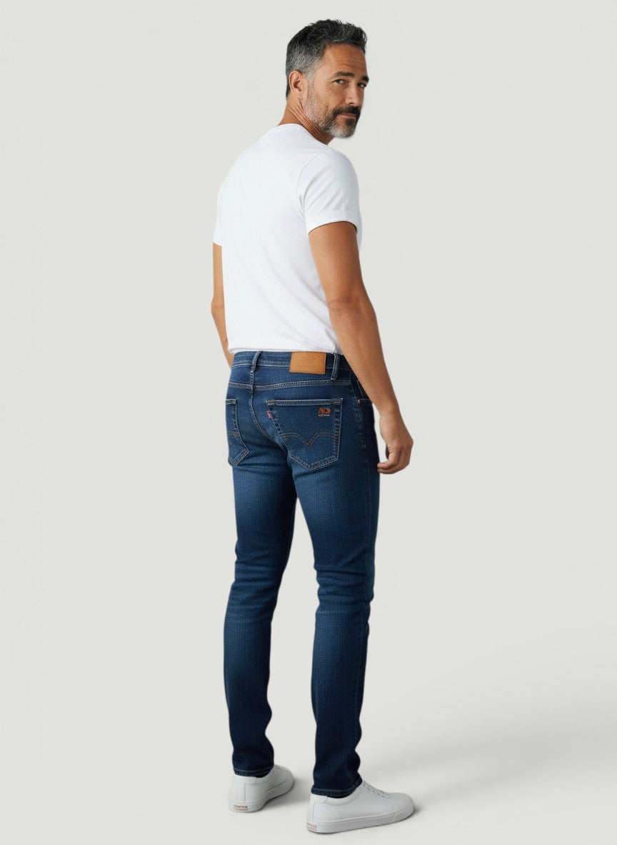 Homem vestindo calça jeans Bluviso azul de corte ajustado, vista traseira, combinada com camiseta branca e tênis.
