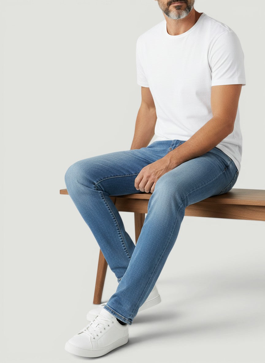 Homem sentado vestindo calça jeans Bluviso azul de corte ajustado com camiseta branca e tênis casual