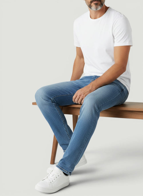 Homem sentado vestindo calça jeans Bluviso azul de corte ajustado com camiseta branca e tênis casual