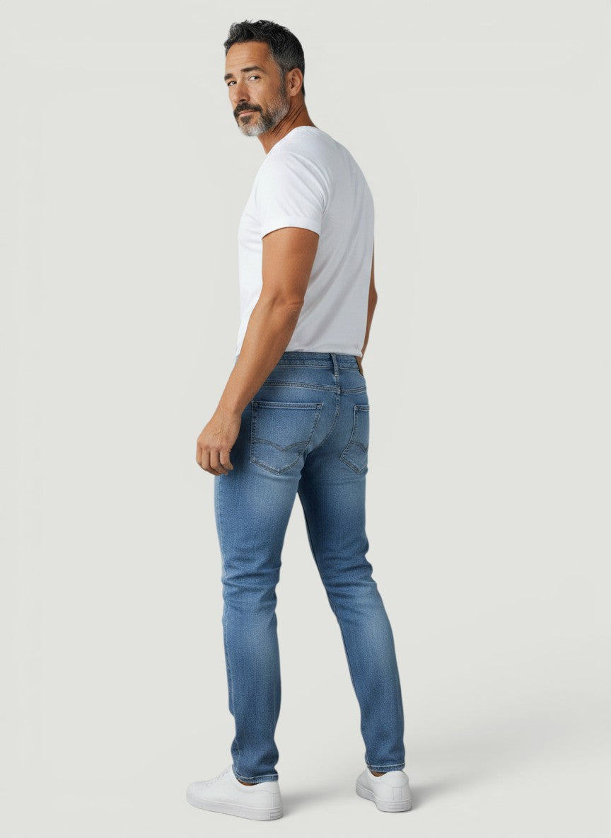 Homem vestindo calça jeans Bluviso azul de corte ajustado com camiseta branca e tênis em fundo neutro