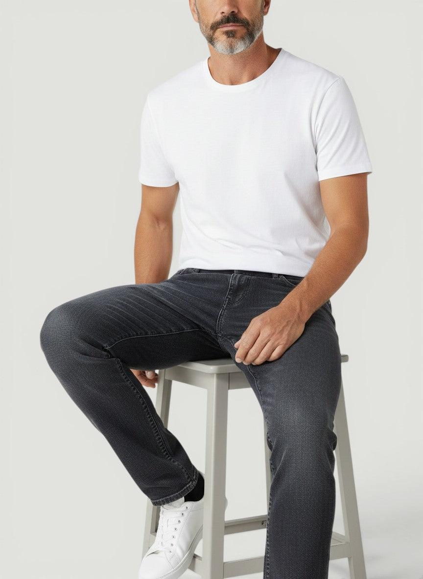 Homem sentado vestindo calça jeans Bluviso cinza de corte ajustado com camiseta branca e tênis casual