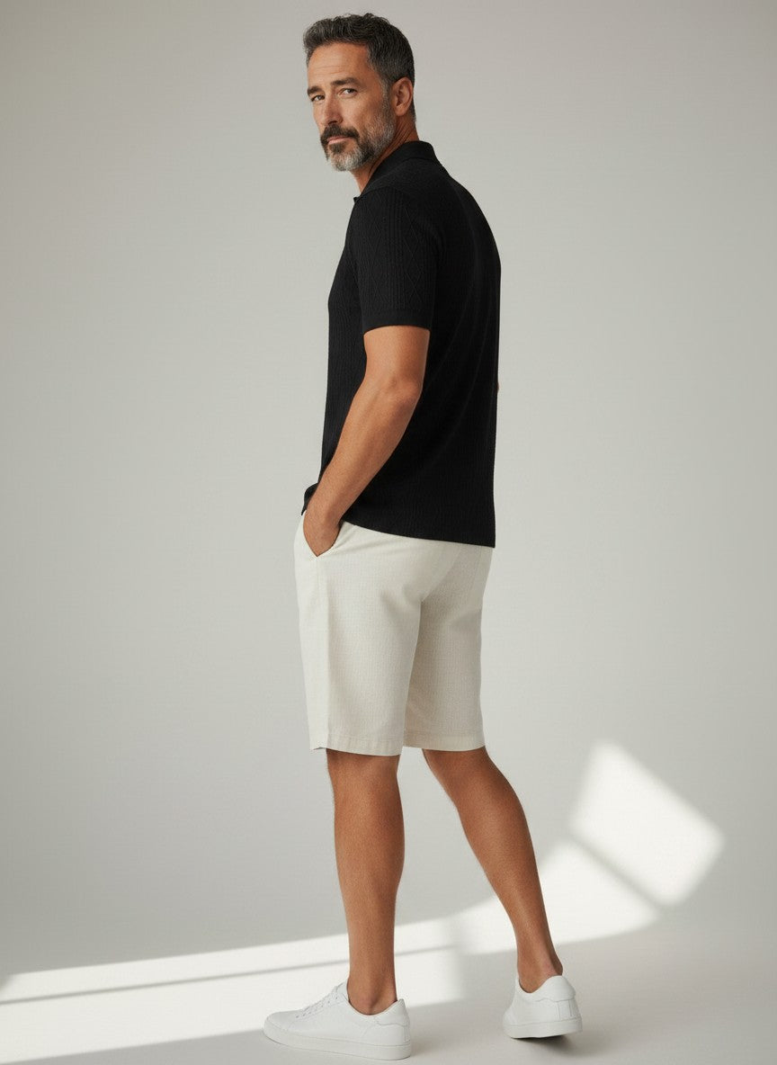 Homem vestindo camisa polo preta texturizada Bluviso com bermuda bege e tênis branco em fundo neutro
