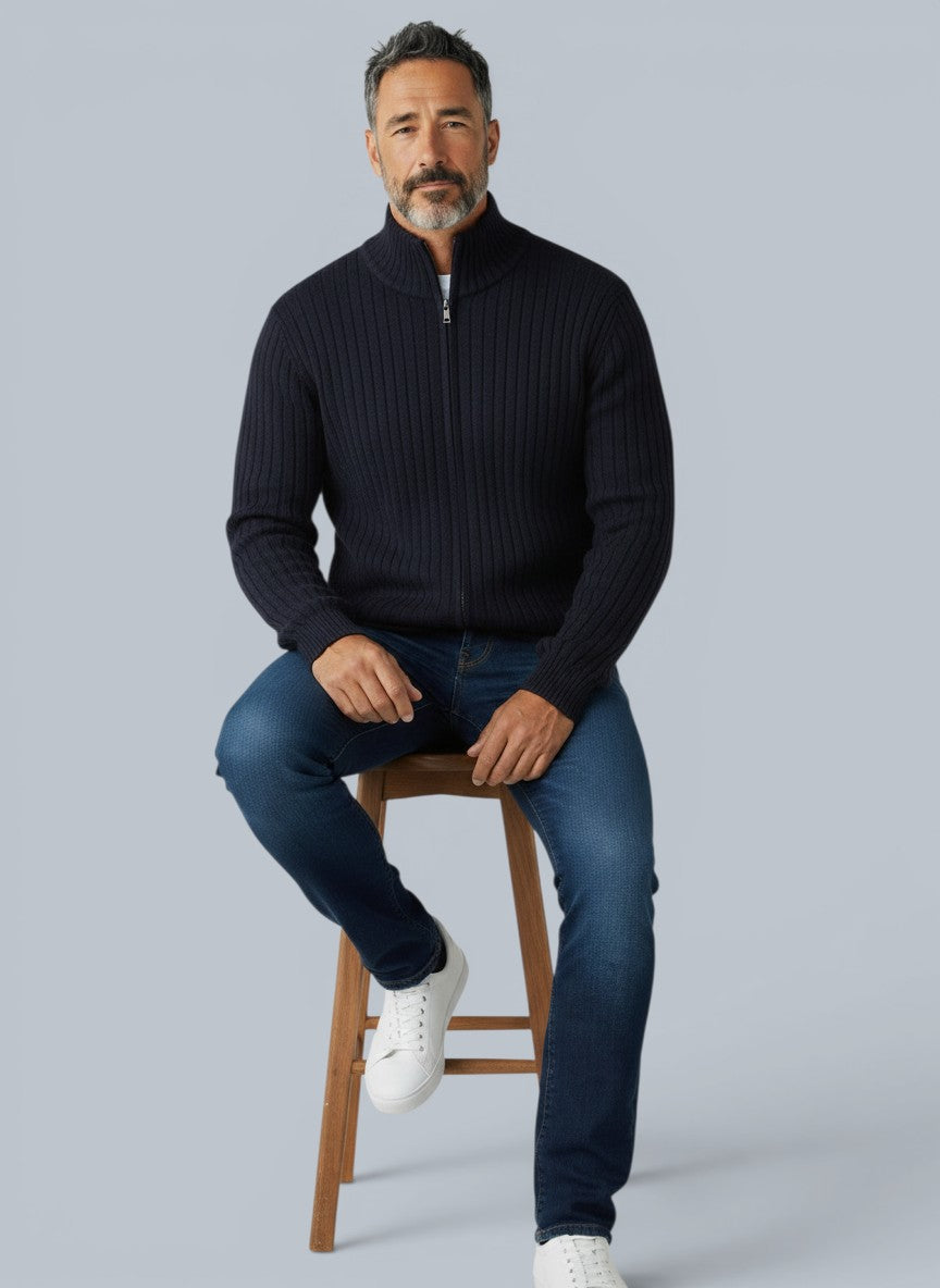 Homem vestindo cardigã de lã merino azul marinho com zíper e gola alta da marca Bluviso, sentado em um banco de madeira.