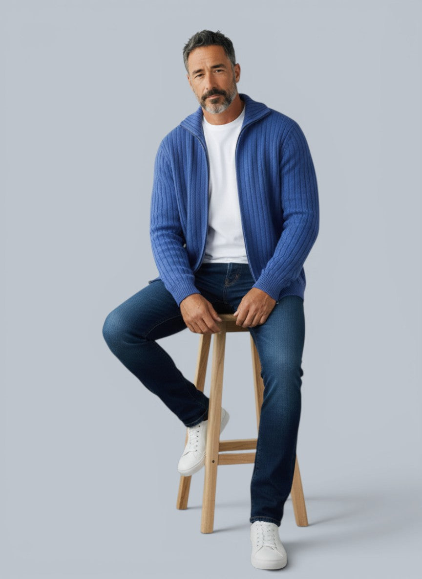 Homem vestindo cardigã de lã merino azul claro com zíper da marca Bluviso, sentado em um banco de madeira em estúdio.